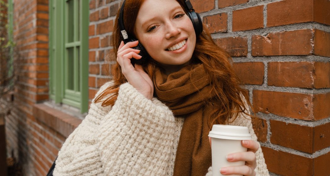 A person with red hair is wearing headphones and a scarf while holding a coffee cup, standing outside near a brick wall.