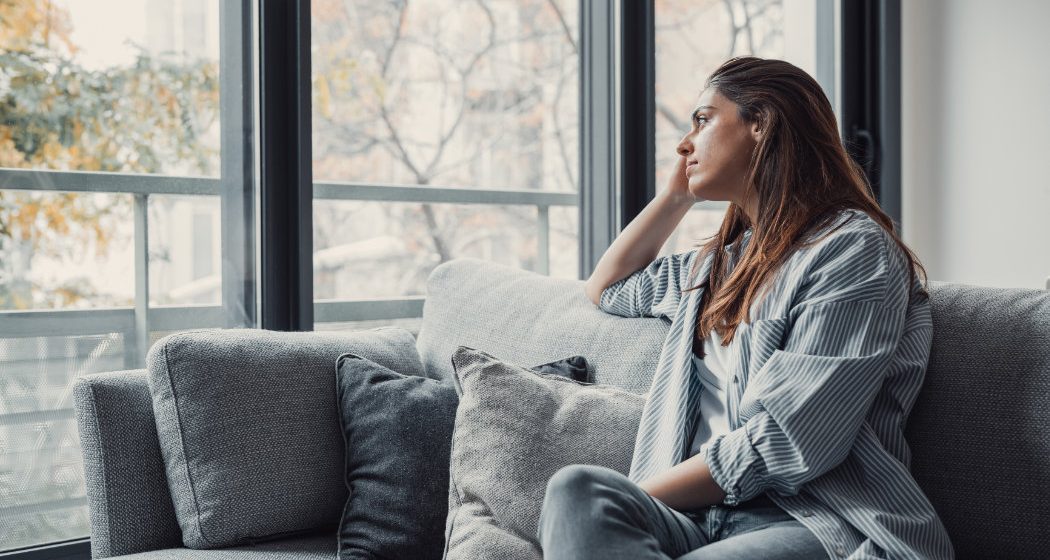Woman sitting on a couch, looking pensively out a large window. She rests her chin on her hand, with trees visible outside.