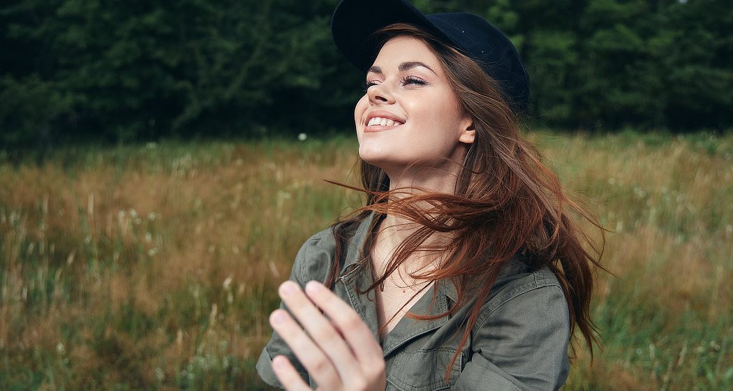 A woman in a green shirt and black cap smiles while standing in a grassy field with trees in the background.