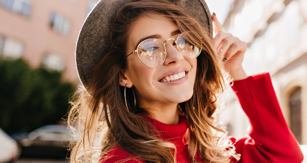 Woman in a red sweater smiles while touching her hat. She is wearing round glasses and hoop earrings, standing outdoors with buildings in the background.