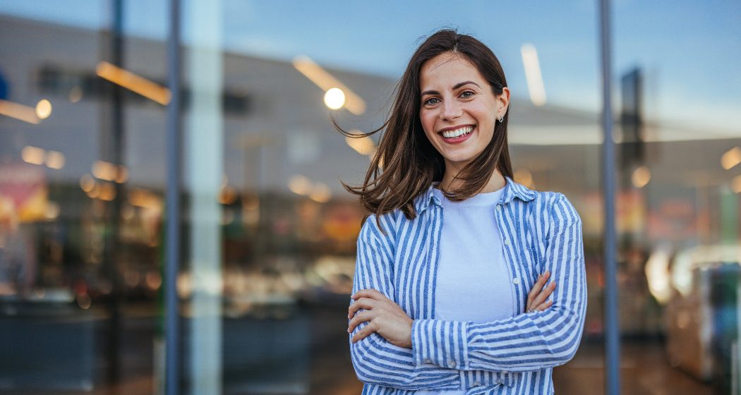 A woman with long brown hair smiles with arms crossed, standing in front of a glass building entrance. She is wearing a white shirt and a blue-and-white striped shirt.