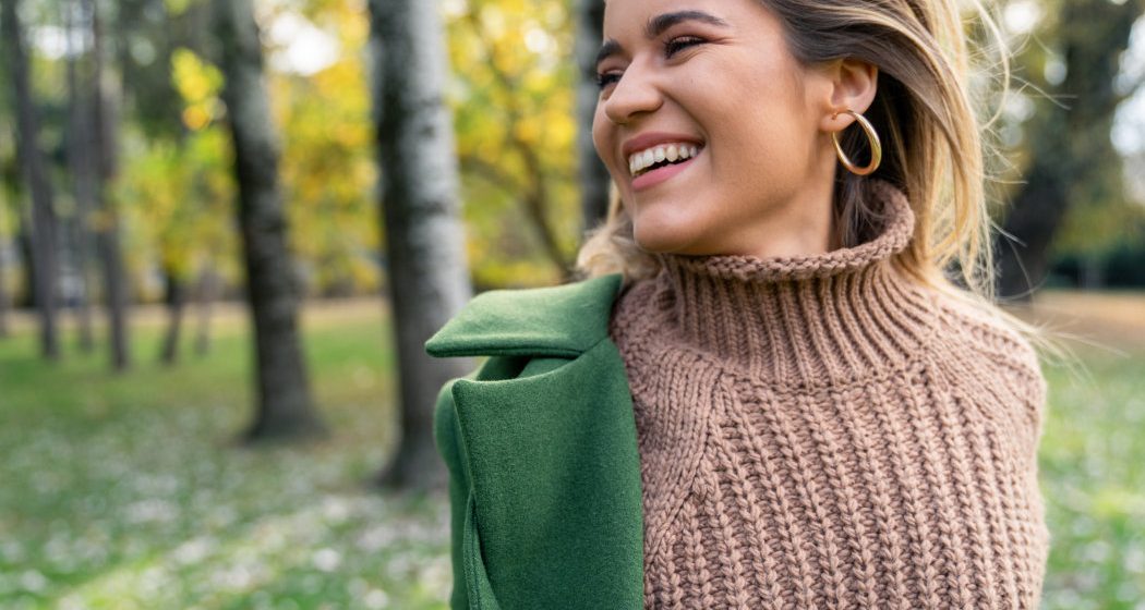 A person wearing a brown knit sweater and green coat stands outdoors, smiling, with blurred trees and grass in the background.