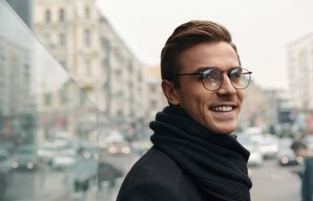 Man wearing glasses and a black scarf smiles while standing outdoors in an urban setting.