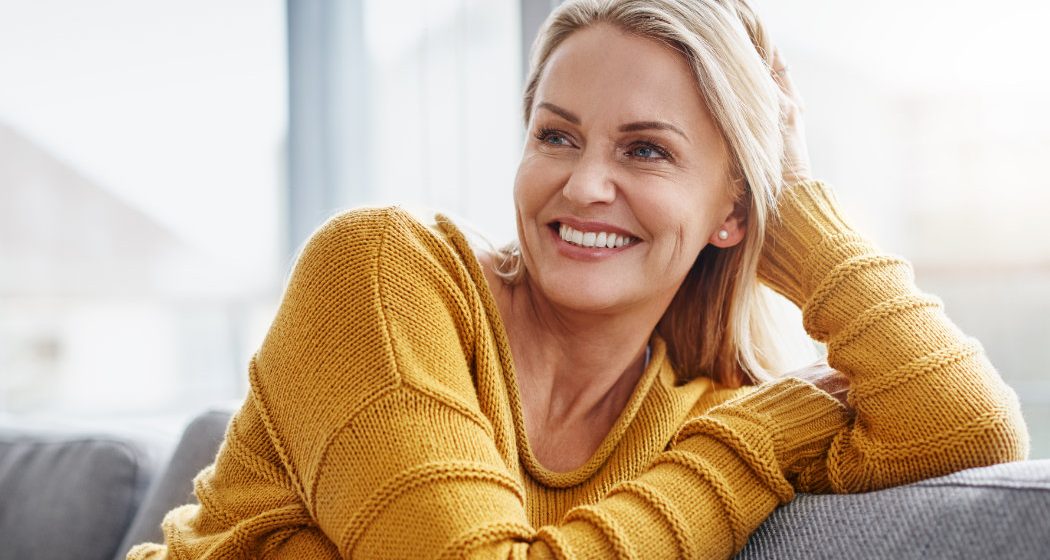 A woman in a yellow sweater sits on a couch, smiling and looking to her left with her hand resting on her head.